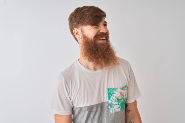 Молодий рудий ірландський чоловік у футболці стоїть над ізольованим білим тлом, дивлячись в сторону з посмішкою на обличчі, природним виразом. Сміється впевнено
. - Фото, зображення