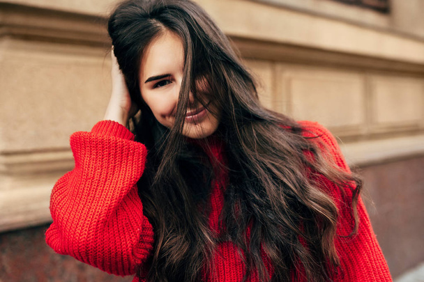 Smiling European brunette woman with long hair looking directly to the camera. Outdoor portrait of blissful female model in trendy knitted red sweater posing during walking in the city street. - Foto, Imagen