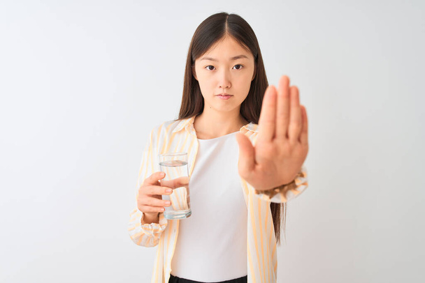 Молодая китаянка в очках пьет стакан воды на изолированном белом фоне с открытой рукой делает знак стоп с серьезным и уверенным выражением, защитный жест
 - Фото, изображение