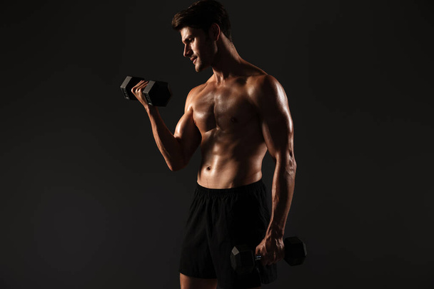 Concentrated handsome young strong sportsman posing isolated over black wall background holding dumbbells. - 写真・画像