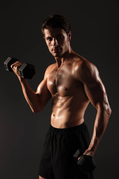 Concentrated handsome young strong sportsman posing isolated over black wall background holding dumbbells. - Фото, зображення