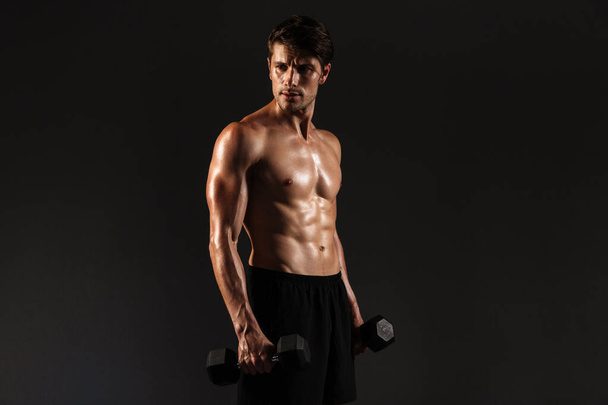 Concentrated handsome young strong sportsman posing isolated over black wall background holding dumbbells. - Zdjęcie, obraz