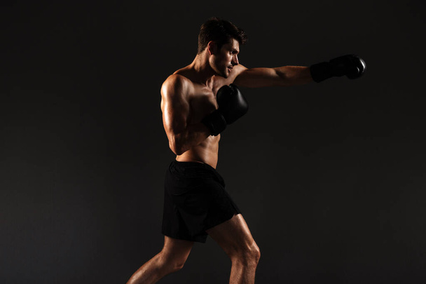 Красивый молодой сильный спортсмен боксер в перчатках делают упражнения бокса изолированы на фоне черной стены
. - Фото, изображение