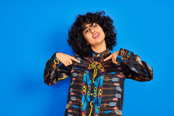 Mladý arabky žena s kudrnaté vlasy na sobě barevné tričko přes izolované modré pozadí vypadá sebevědomě s úsměvem na tváři, ukazuje sám s prsty hrdý a šťastný. - Fotografie, Obrázek