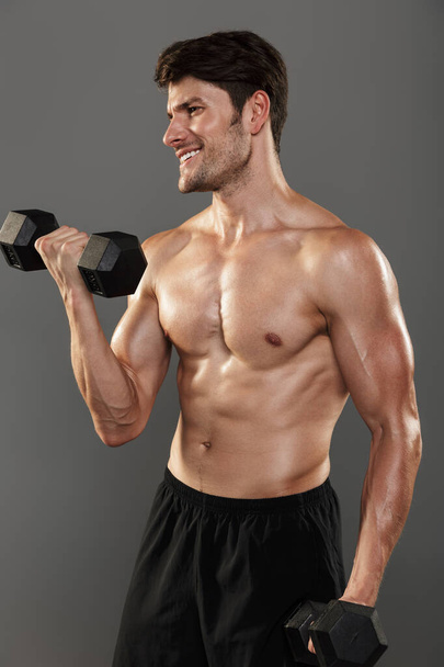 Cheerful happy handsome young strong sportsman posing isolated over grey wall background make exercises with dumbbells for arms. - Foto, Imagem