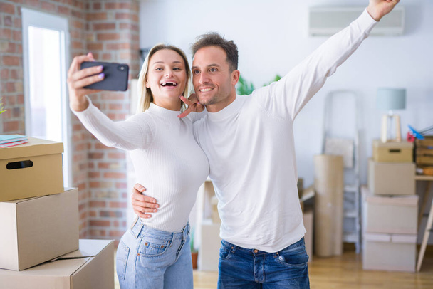 Joven hermosa pareja de pie utilizando el teléfono inteligente para tomar selfie en el nuevo hogar alrededor de cajas de cartón
 - Foto, imagen