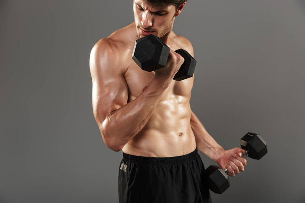 Handsome young strong sportsman posing isolated over grey wall background make exercises with dumbbells for arms. - Photo, Image