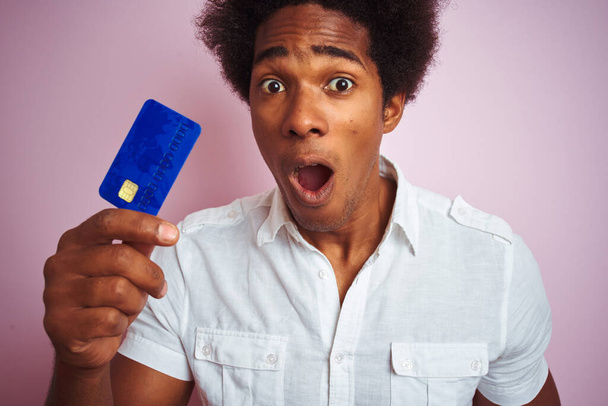 Afro amerikai ügyfél férfi kezében hitelkártya felett áll elszigetelt rózsaszín háttér megijedt a sokk egy meglepetés arc, fél, és izgatott a félelem kifejezés - Fotó, kép