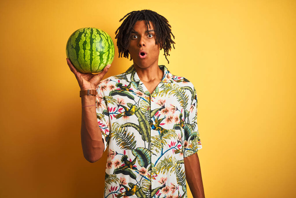 Afro-Amerikaanse man met dreadlocks houden watermeloen over geïsoleerde gele achtergrond bang in shock met een verrassing gezicht, bang en opgewonden met angst uitdrukking - Foto, afbeelding