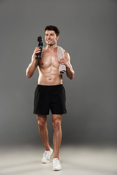 Handsome young strong sportsman isolated over grey wall background holding towel and bottle with water. - Foto, Imagen