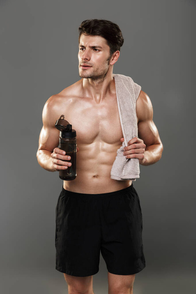 Handsome young strong sportsman isolated over grey wall background holding towel and bottle with water. - Fotoğraf, Görsel