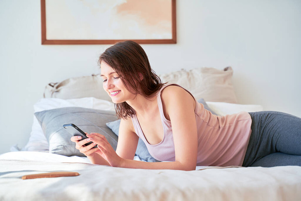 Photo of young brunette with phone in her hands lying on bed - Photo, Image
