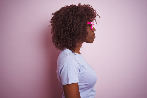 Молодая африканская женщина в футболках очки на изолированном розовом фоне глядя в сторону, расслабиться профиль позировать с естественным лицом с уверенной улыбкой
. - Фото, изображение