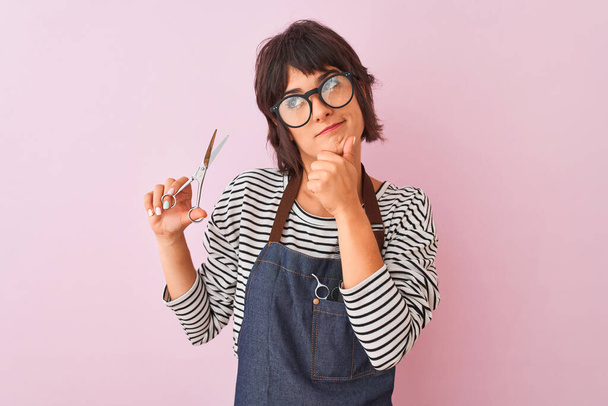 Hairdresser woman wearing apron and glasses holding scissors over isolated pink background serious face thinking about question, very confused idea - 写真・画像