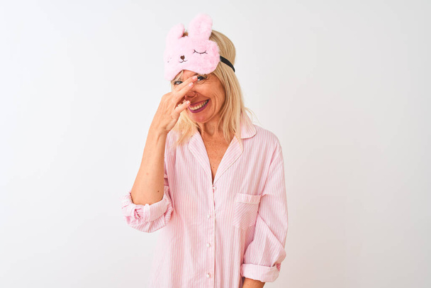 Middle age woman wearing sleep mask and pajama over isolated white background smelling something stinky and disgusting, intolerable smell, holding breath with fingers on nose. Bad smells concept. - Fotoğraf, Görsel