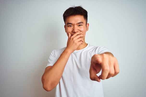 Young asian chinese man wearing t-shirt standing over isolated white background laughing at you, pointing finger to the camera with hand over mouth, shame expression - 写真・画像