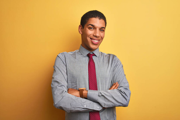 Νεαρός όμορφος Άραβας επιχειρηματίας φορώντας πουκάμισο και γραβάτα πάνω από απομονωμένο κίτρινο φόντο χαρούμενο πρόσωπο χαμογελώντας με σταυρωμένα χέρια κοιτάζοντας την κάμερα. Θετικό πρόσωπο. - Φωτογραφία, εικόνα