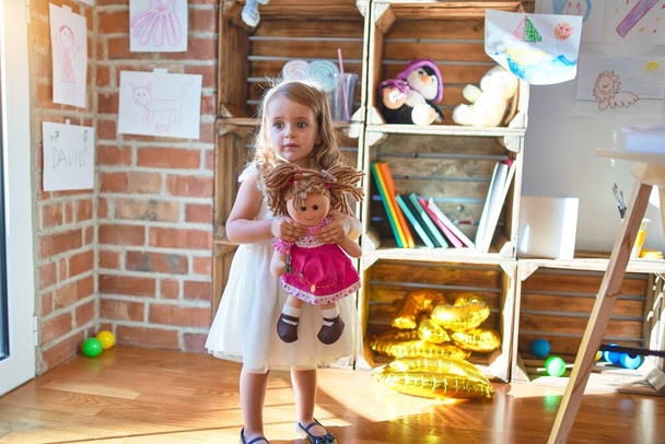 Очаровательная блондинка обнимает куклу вокруг большого количества игрушек в детском саду
 - Фото, изображение