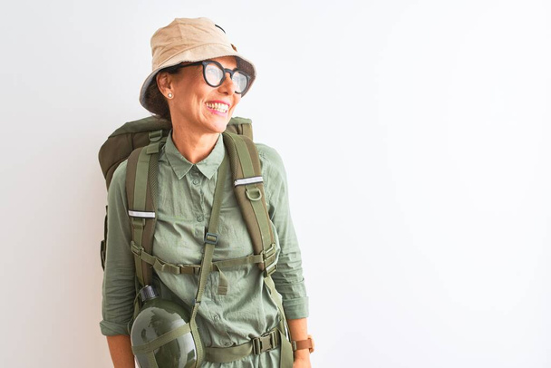 Middle age hiker woman wearing backpack canteen hat glasses over isolated white background looking away to side with smile on face, natural expression. Laughing confident. - Zdjęcie, obraz