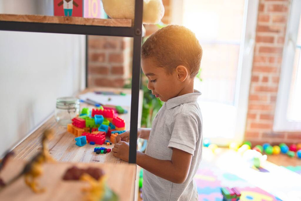 Hermoso niño afroamericano de pie jugando con pequeños bloques de construcción en estanterías en el jardín de infantes
 - Foto, imagen