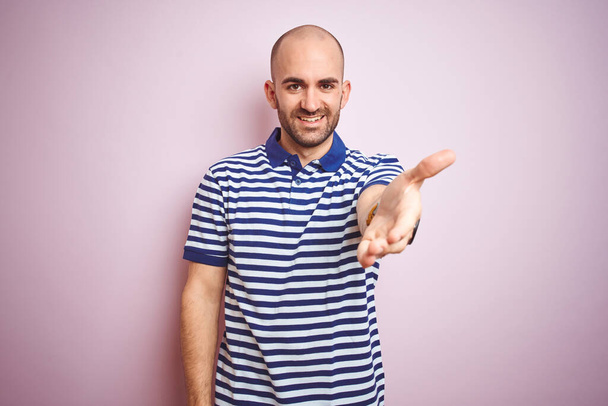 Nuori kalju mies, jolla on parta yllään rento raidallinen sininen t-paita vaaleanpunainen eristetty tausta hymyilevä ystävällinen tarjoamalla kädenpuristus tervehdys ja kutsuva. Menestyksekäs yritys
. - Valokuva, kuva