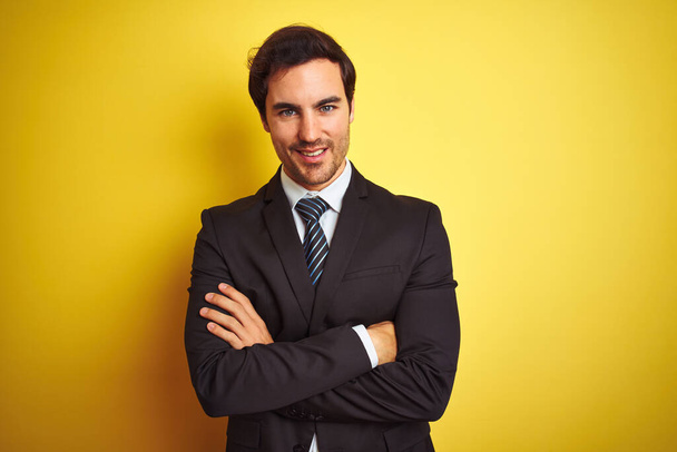 Young handsome businessman wearing suit and tie standing over isolated yellow background happy face smiling with crossed arms looking at the camera. Positive person. - Photo, Image