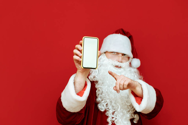 Santa Claus stands with smartphone in hand on red background and shows finger on blank white screen. Focus on smartphone screen. Isolated. Copyspace - Φωτογραφία, εικόνα