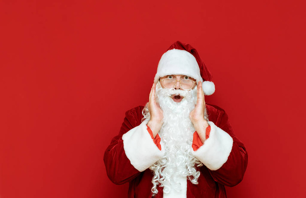 Astonished Santa Claus stands against a red wall and looks into the camera. Portrait of shocked man in santa costume isolated. Santa was surprised at the discounts for Christmas. Copyspace - Fotografie, Obrázek