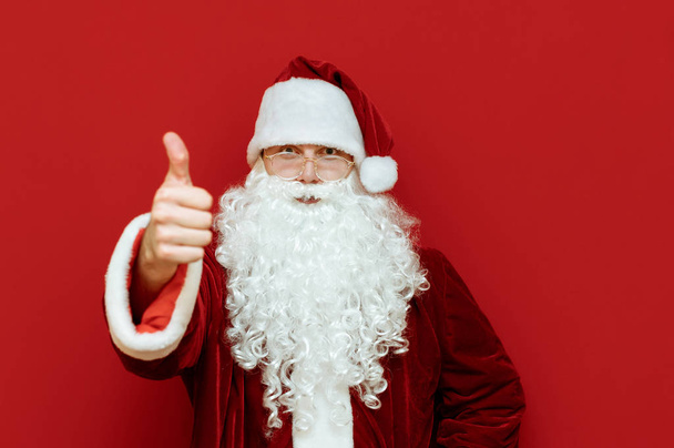 Portrait of beautiful Santa Claus in red suit standing on red background and showing thumb up. Happy Santa. Isolated. Christmas. Copyspace - Fotografie, Obrázek