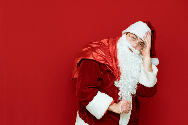Tired Santa Claus with a bag on his back stands on a red background and looks into the camera, Santa is tired of carrying presents. Delivery of Christmas gifts by Santa Claus. Xmas concept - Photo, Image