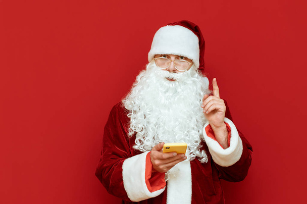 Portrait of Santa Claus on red background with smartphone in hand stands on red background, looks into camera with serious face and raised forefinger. Christmas concept. Copyspace - Fotó, kép