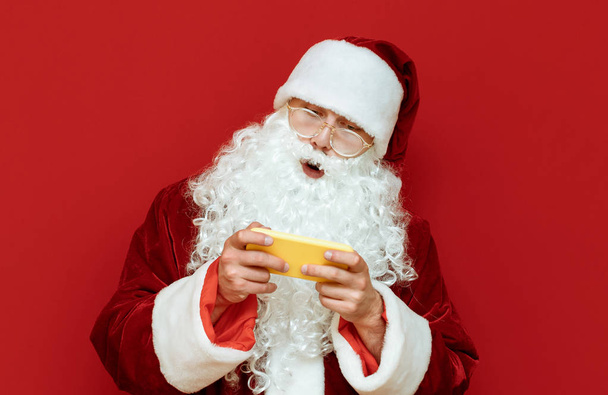 Santa Claus jugador juega videojuegos en el teléfono inteligente, se ve sorprendido en la pantalla aislada en el fondo rojo. Retrato de santa gamer. Juegos de Navidad
. - Foto, imagen