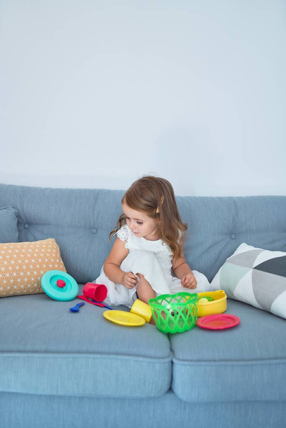 Adorable blond tout-petit assis sur le canapé cuisine avec de la nourriture et des couverts jouets à la maison
 - Photo, image