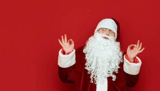 Retrato de Santa Claus satisfecho con los ojos cerrados muestra gesto de las manos Ok aislado sobre fondo rojo. Copyspace a la derecha. Retrato de santa feliz meditando sobre fondo rojo. X mas
 - Foto, Imagen