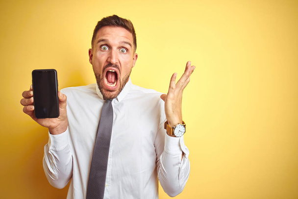 Young handsome business man showing smartphone screen over yellow isolated background very happy and excited, winner expression celebrating victory screaming with big smile and raised hands - Photo, Image