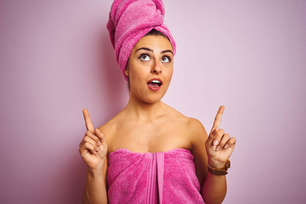 Jovem mulher bonita vestindo toalha após o chuveiro sobre fundo rosa isolado espantado e surpreso olhando para cima e apontando com os dedos e braços levantados
. - Foto, Imagem