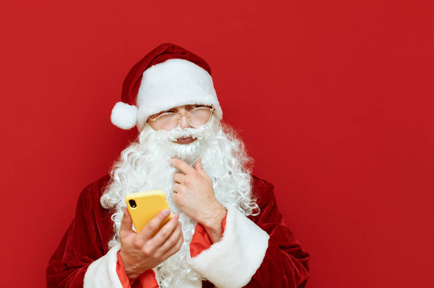 Studio photo of funny Santa with smartphone in hand on red background, looking away at blank space with pensive face. Santa with Phone and Copyspace. X-mas concept - Photo, Image