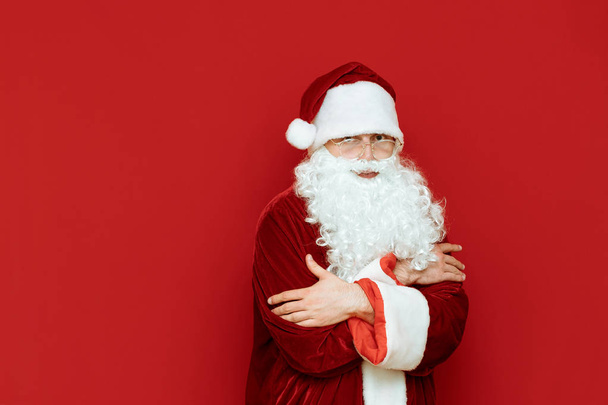 Санта-Клаус замерзает, согревается на красном фоне и смотрит в камеру с грустным лицом. Человек в костюме Санты замерзает на красном фоне. Холодная зима на Рождество
 - Фото, изображение