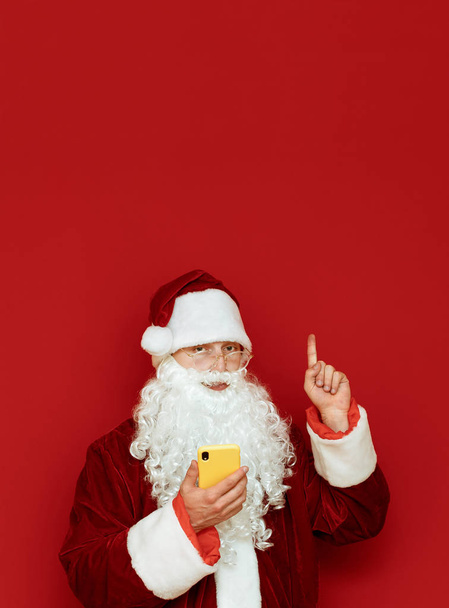 Άγιος Βασίλης στέκεται σε κόκκινο φόντο με smartphone στο χέρι, κοιτάζει στην κάμερα με σοβαρό πρόσωπο και δείχνει τον αντίχειρα επάνω στο copyspace. Απομονωμένο πορτρέτο. Κάθετη φωτογραφία. - Φωτογραφία, εικόνα