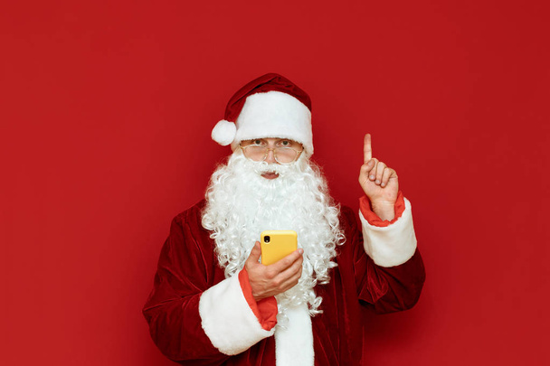 Portrait of a funny Santa Claus standing on a red background with a smartphone in his hand, looking into the camera and pointing his finger up at an empty space copyspace. X-mas - Photo, Image