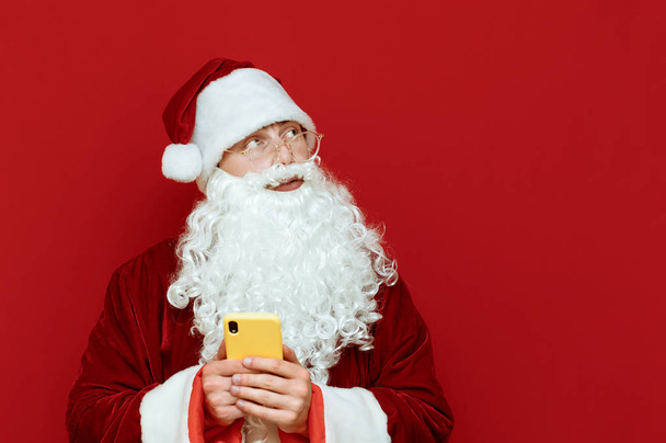 Elinde akıllı telefonla kırmızı arka planda duran ve boş telif alanına bakan dalgın Noel Baba 'nın portresi. Noel Baba düşünceli bir şekilde kırmızıda izole edilmiş. Noel ve Yeni Yıl kavramı. - Fotoğraf, Görsel