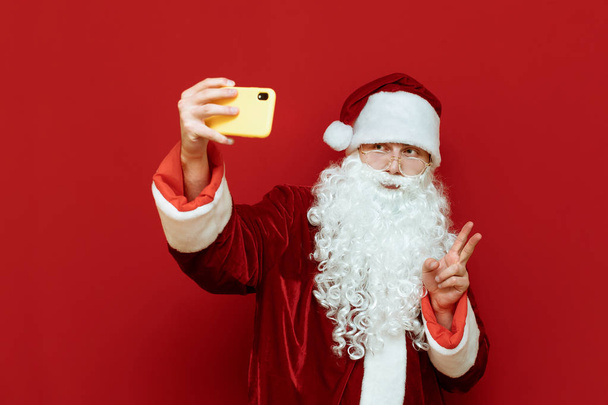 Studio φωτογραφία του Αϊ Βασίλη ποζάρουν σε κάμερα smartphone, λαμβάνοντας selfies και δείχνει την ειρήνη χειρονομία στην κάμερα σε κόκκινο φόντο. Σέλφι. Άγιος Βασίλης και τεχνολογία. Καλά Χριστούγεννα.. - Φωτογραφία, εικόνα