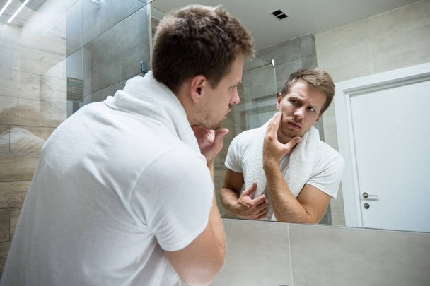 young handsome man with white towel on his shoulders looking in the bathroom mirror touching his face - Фото, зображення