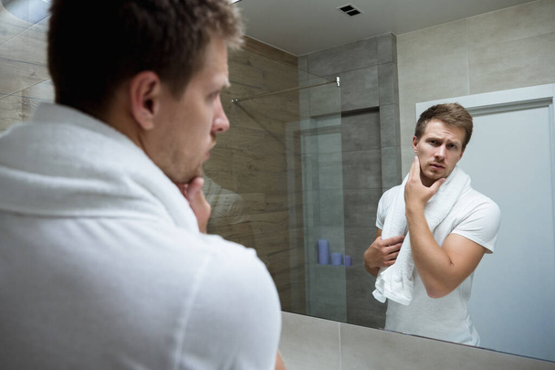 young handsome man with white towel on his shoulders looking in the bathroom mirror touching his face with one hand - Photo, Image