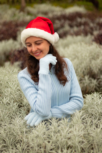 Νεαρή γυναίκα με κόκκινο καπέλο και μπλε γάντια. Όμορφη λευκή γυναίκα χαμογελάει, κοιτάζει κάτω και αγγίζει το πηγούνι της. Χριστούγεννα και Πρωτοχρονιά γιορτή.  - Φωτογραφία, εικόνα