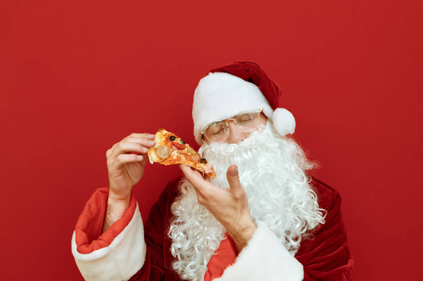 Nahaufnahme Porträt des Weihnachtsmannes mit einem Stück Pizza in den Händen mit geschlossenen Augen auf rotem Hintergrund, isoliert. Weihnachtsmann in rotem Anzug, Bart und Brille posiert mit einem Stück Pizza. - Foto, Bild