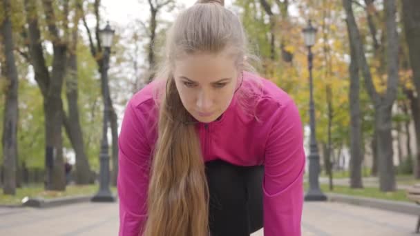 Close-up of confident young Caucasian girl at start position looking up and starting running. Attractive female runner training in the morning in the autumn park. Camera zooms in. - Footage, Video
