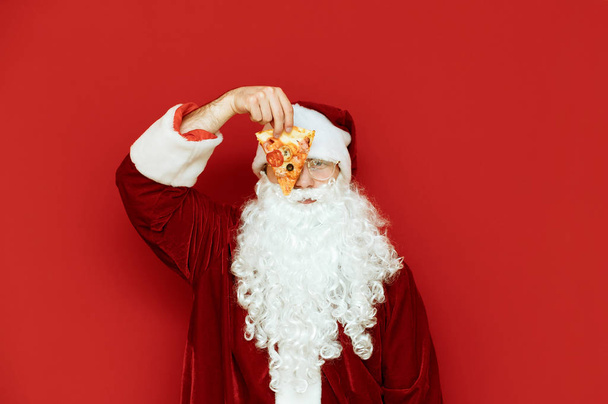 Zabawny człowiek w kostiumie Świętego Mikołaja stoi na czerwonym tle, kładzie kawałek pizzy na oku, patrzy w kamerę i się śmieje. Zabawny portret Mikołaja z plasterkiem pizzy.Boże Narodzenie, koncepcja Nowego Roku - Zdjęcie, obraz
