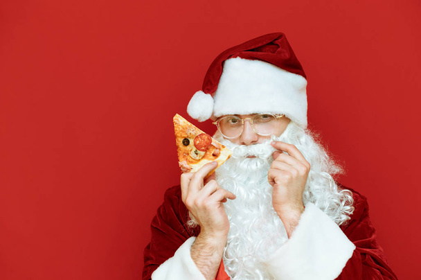 Lustiger Weihnachtsmann im roten Kostüm steht mit Pizzastück auf rotem Grund, blickt in die Kamera und streicht sich über den Schnurrbart. Pizza zu Weihnachten. Der Weihnachtsmann liebt Pizza. Leerraum, um auf der linken Seite Werbung zu machen. isoliert - Foto, Bild