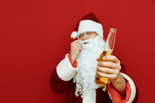 Eine Flasche Bier in der Hand des Weihnachtsmannes steht auf rotem Grund, lächelt und kratzt sich mit der Hand an seinem Schnurrbart. konzentrieren sich auf die Hand des Weihnachtsmannes mit einer Flasche Bier. Vereinzelt. Weihnachtsmann und Alkohol - Foto, Bild